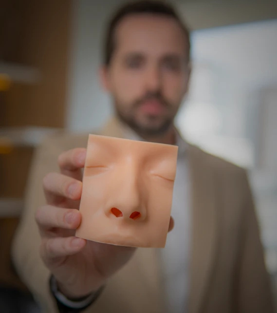 Dr, Thiago Chequetto com o rosto desfocado ao fundo, segura à frente da câmera um modelo de silicone de nariz e olhos usado para demonstrar cirurgia nasal.